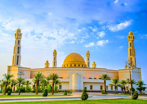 A large mosque featuring a central golden dome flanked by two tall minarets. The building is accented with arches and surrounded by palm trees, under a clear blue sky with scattered clouds.