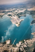 An aerial view of a large industrial port located along a coastline with clear blue waters. The port is surrounded by various infrastructure elements including warehouses, roads, and storage tanks. A few ships are visible in the water near the docks, and there are areas of green vegetation scattered throughout the landscape.