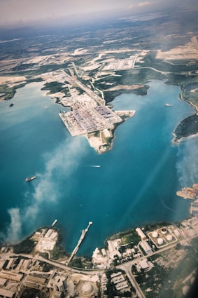An aerial view of a large industrial port located along a coastline with clear blue waters. The port is surrounded by various infrastructure elements including warehouses, roads, and storage tanks. A few ships are visible in the water near the docks, and there are areas of green vegetation scattered throughout the landscape.