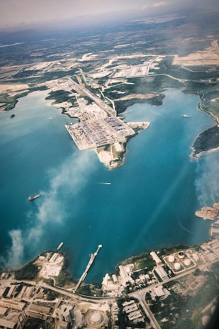 An aerial view of a large industrial port located along a coastline with clear blue waters. The port is surrounded by various infrastructure elements including warehouses, roads, and storage tanks. A few ships are visible in the water near the docks, and there are areas of green vegetation scattered throughout the landscape.
