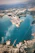 An aerial view of a large industrial port located along a coastline with clear blue waters. The port is surrounded by various infrastructure elements including warehouses, roads, and storage tanks. A few ships are visible in the water near the docks, and there are areas of green vegetation scattered throughout the landscape.