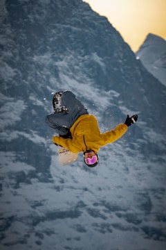 A snowboarder performs an aerial trick against a backdrop of snowy mountains, wearing a yellow jacket and grey pants. The snowboarder's goggles are a reflective pink color, highlighting the dynamic motion as snowflakes are visible around them.