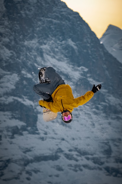 A snowboarder performs an aerial trick against a backdrop of snowy mountains, wearing a yellow jacket and grey pants. The snowboarder's goggles are a reflective pink color, highlighting the dynamic motion as snowflakes are visible around them.
