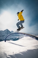 A snowboarder wearing a yellow jacket and a helmet is performing a trick on a curved rail against a backdrop of snowy mountains and a bright sky. The person is in mid-air, showcasing an action-packed moment.