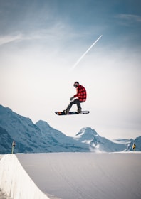 A snowboarder in mid-air performing a jump against a backdrop of snow-covered mountains. The snowboarder is wearing a red and black checkered jacket and helmet, while holding a snowboard. The sky is clear with a visible contrail from an airplane.