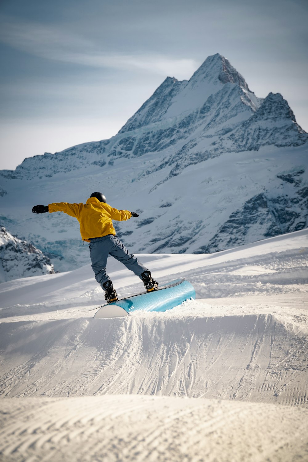 黄色いジャケットと青いズボンを着た男性が、昼間、雪に覆われた山で青いスノーボードに乗っています