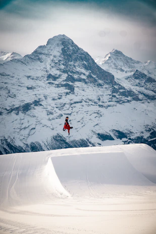 A dynamic snowscoot rider launching off a snowy mountain jump with Crans-Montana peaks in the background.
