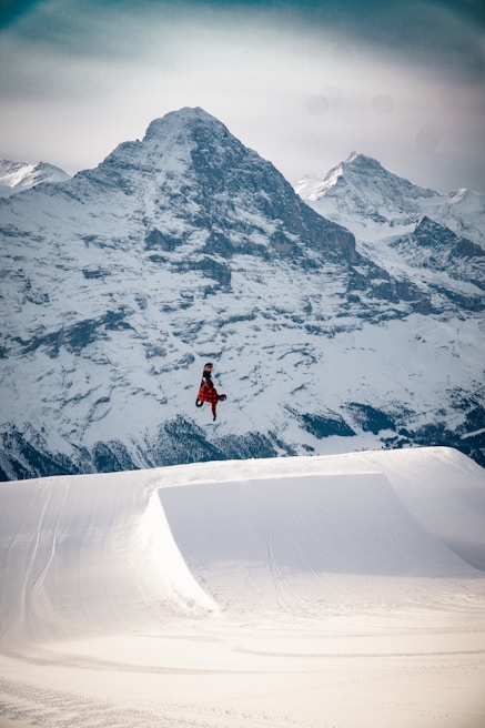 A snowboarder in mid-air performing a trick against a backdrop of majestic snow-covered mountains. The snowboarder is wearing a red and black checkered outfit, emphasizing the action against the white snow. The large snow ramp in the foreground highlights the scale of the jump.