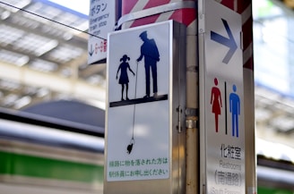 A sign at a train station depicts a silhouette of an adult and a child holding hands, with a dropped object suspended on a fishing line, possibly indicating safety instructions. The sign is alongside another indicating restrooms, with symbols for male and female. The background shows blurred structures typical of a train platform.