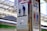 A sign at a train station depicts a silhouette of an adult and a child holding hands, with a dropped object suspended on a fishing line, possibly indicating safety instructions. The sign is alongside another indicating restrooms, with symbols for male and female. The background shows blurred structures typical of a train platform.