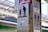 A sign at a train station depicts a silhouette of an adult and a child holding hands, with a dropped object suspended on a fishing line, possibly indicating safety instructions. The sign is alongside another indicating restrooms, with symbols for male and female. The background shows blurred structures typical of a train platform.