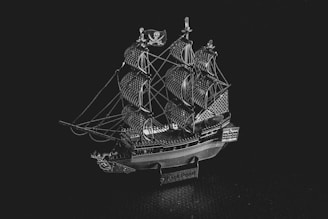 A detailed miniature model of a classic pirate ship with multiple sails, intricate rigging, and a small flag featuring a skull emblem. The model is metallic and displays fine craftsmanship.