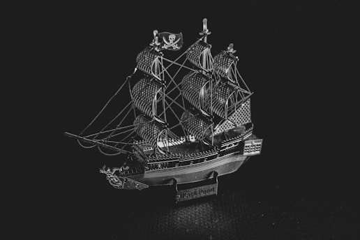 A detailed miniature model of a classic pirate ship with multiple sails, intricate rigging, and a small flag featuring a skull emblem. The model is metallic and displays fine craftsmanship.
