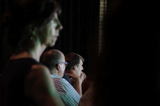 A darkened room with several people appearing to be listening intently. The scene is dimly lit, with one person in the foreground out of focus, and others slightly clearer in the midground, suggesting an indoor event or presentation.