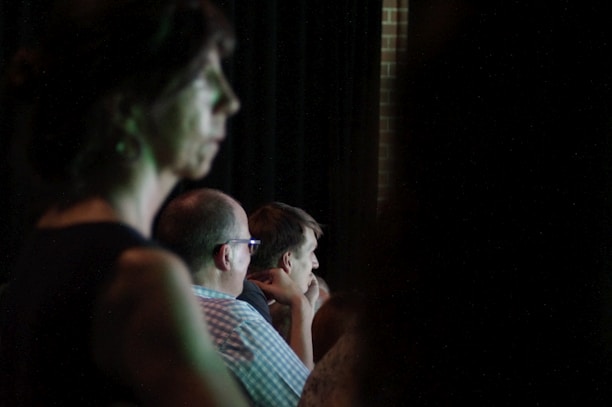 A darkened room with several people appearing to be listening intently. The scene is dimly lit, with one person in the foreground out of focus, and others slightly clearer in the midground, suggesting an indoor event or presentation.
