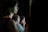 A darkened room with several people appearing to be listening intently. The scene is dimly lit, with one person in the foreground out of focus, and others slightly clearer in the midground, suggesting an indoor event or presentation.