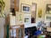 An artist's workspace with an easel holding a painting of a bird perched on a faucet. Multiple paintings hang on a wall, featuring nature scenes and abstract designs. The room is filled with art supplies, including brushes, canvases, and tools scattered around the area.