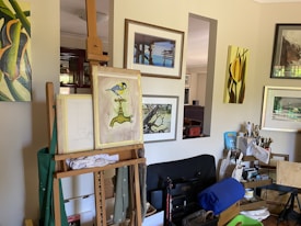 An artist's workspace with an easel holding a painting of a bird perched on a faucet. Multiple paintings hang on a wall, featuring nature scenes and abstract designs. The room is filled with art supplies, including brushes, canvases, and tools scattered around the area.