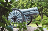 A historic cannon is situated in a lush green park setting, surrounded by vibrant foliage. The cannon is made of metal with visible rivets and wheels, resting on a stone platform. Trees and manicured grass form the background, contributing to a serene and preserved atmosphere.
