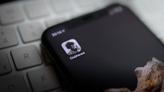 A smartphone displays the Clubhouse app icon on its screen, lying on top of a computer keyboard. The screen is mostly black with a monochrome logo, showing a high-contrast image of a person. Part of a wooden textured phone case is visible at the bottom.