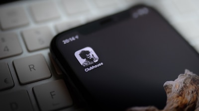 A smartphone displays the Clubhouse app icon on its screen, lying on top of a computer keyboard. The screen is mostly black with a monochrome logo, showing a high-contrast image of a person. Part of a wooden textured phone case is visible at the bottom.