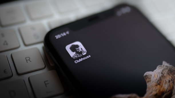 A smartphone displays the Clubhouse app icon on its screen, lying on top of a computer keyboard. The screen is mostly black with a monochrome logo, showing a high-contrast image of a person. Part of a wooden textured phone case is visible at the bottom.