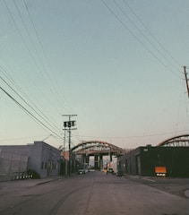 An urban street scene with industrial buildings on both sides and power lines crossing overhead. In the distance, a large steel structure or bridge under construction dominates the view, with some parked vehicles along the roadside.