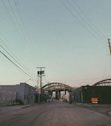 An urban street scene with industrial buildings on both sides and power lines crossing overhead. In the distance, a large steel structure or bridge under construction dominates the view, with some parked vehicles along the roadside.