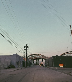 An urban street scene with industrial buildings on both sides and power lines crossing overhead. In the distance, a large steel structure or bridge under construction dominates the view, with some parked vehicles along the roadside.