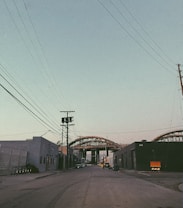 An urban street scene with industrial buildings on both sides and power lines crossing overhead. In the distance, a large steel structure or bridge under construction dominates the view, with some parked vehicles along the roadside.