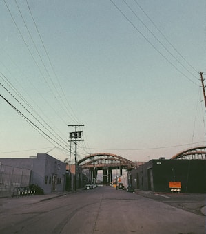 An urban street scene with industrial buildings on both sides and power lines crossing overhead. In the distance, a large steel structure or bridge under construction dominates the view, with some parked vehicles along the roadside.