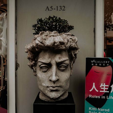 A sculpture of a classical male head with a plant growing from the top is positioned on a pedestal. The stone-like texture gives the figure an ancient, weathered look. Next to the sculpture is a colorful poster with text in both English and Chinese, advertising an art gallery exhibit.
