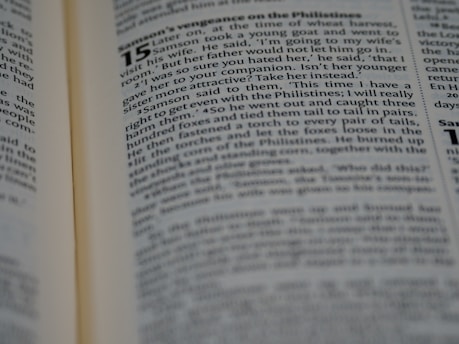 A close-up of an open book with text visible on the right-hand page. The text is written in a structured format, with chapter and verse numbers indicating it is a religious text. The left page is slightly out of focus.