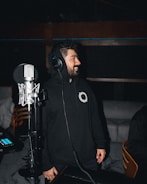 A person wearing a black hoodie with a white circular logo is standing in a dimly lit room, likely a recording studio. The individual is wearing headphones and is positioned next to a professional microphone setup, looking off to the side with a smile.
