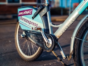 A close-up view of a rental bicycle, prominently displaying the branding 'Durstexpress.de'. The bike has a light blue frame with visible logos and text decals, including 'DING DONG!' and 'nextbike'. The bicycle is parked on a paved street with a blurred urban background.