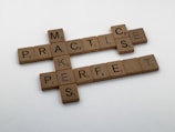 Wooden Scrabble tiles are arranged on a white surface to form the phrase 'Practice Makes Perfect.' Each tile has a letter and a point value, typical of the Scrabble game.