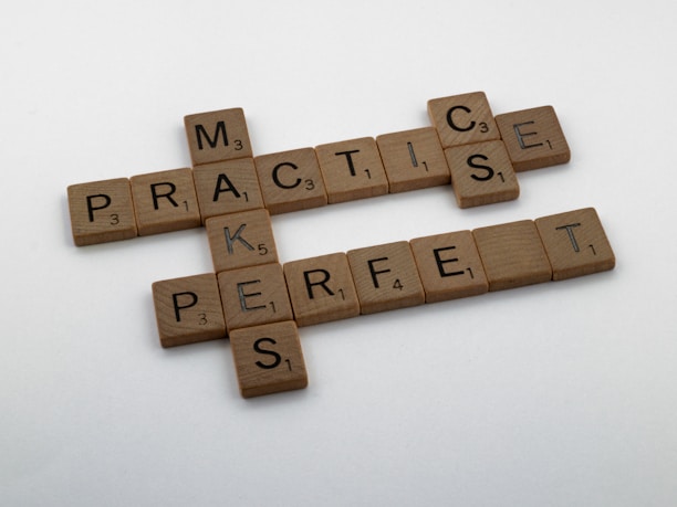 Wooden Scrabble tiles are arranged on a white surface to form the phrase 'Practice Makes Perfect.' Each tile has a letter and a point value, typical of the Scrabble game.