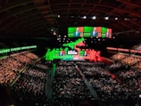 A large indoor conference venue filled with a seated audience. The stage is adorned with brightly colored digital displays in green and red, featuring the words 'Web Summit'. Multiple large screens project an image of a speaker addressing the crowd from the stage.