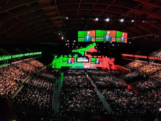 A large indoor conference venue filled with a seated audience. The stage is adorned with brightly colored digital displays in green and red, featuring the words 'Web Summit'. Multiple large screens project an image of a speaker addressing the crowd from the stage.