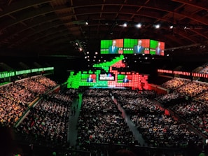 A large indoor conference venue filled with a seated audience. The stage is adorned with brightly colored digital displays in green and red, featuring the words 'Web Summit'. Multiple large screens project an image of a speaker addressing the crowd from the stage.