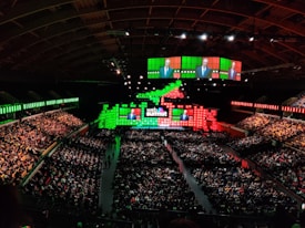 A large indoor conference venue filled with a seated audience. The stage is adorned with brightly colored digital displays in green and red, featuring the words 'Web Summit'. Multiple large screens project an image of a speaker addressing the crowd from the stage.