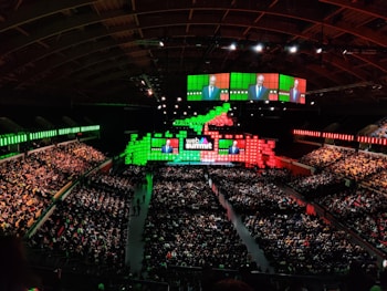 A large indoor conference venue filled with a seated audience. The stage is adorned with brightly colored digital displays in green and red, featuring the words 'Web Summit'. Multiple large screens project an image of a speaker addressing the crowd from the stage.