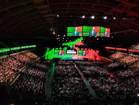 A large indoor conference venue filled with a seated audience. The stage is adorned with brightly colored digital displays in green and red, featuring the words 'Web Summit'. Multiple large screens project an image of a speaker addressing the crowd from the stage.