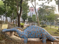 A large blue dinosaur sculpture with brown spots is positioned near a staircase in an outdoor park setting. Surrounding the dinosaur are various trees and greenery, and in the background, a multi-story residential building is visible. The scene appears peaceful with a clear sky and well-maintained surroundings.