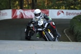 A motorcyclist in full gear is engaged in a race, leaning aggressively into a turn on a winding road surrounded by greenery. The motorcycle features prominent sponsor decals. Another rider follows closely behind, creating a sense of speed and competition.