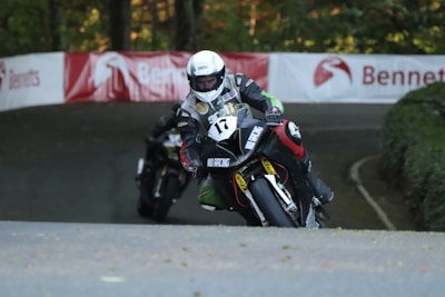 A motorcyclist in full gear is engaged in a race, leaning aggressively into a turn on a winding road surrounded by greenery. The motorcycle features prominent sponsor decals. Another rider follows closely behind, creating a sense of speed and competition.