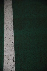 A vertical white line painted on a dark green textured surface, possibly a sports court or a parking lot. The paint appears worn and slightly irregular, with small specks and imperfections visible along the line.