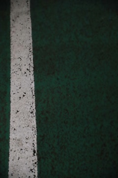 A vertical white line painted on a dark green textured surface, possibly a sports court or a parking lot. The paint appears worn and slightly irregular, with small specks and imperfections visible along the line.