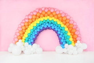 A vibrant arch made of colorful balloons mimicking a rainbow. The arrangement features red, orange, yellow, green, blue, and purple balloons, forming a semicircular shape. At the base of the arch are clusters of white balloons resembling clouds, set against a soft pink background.