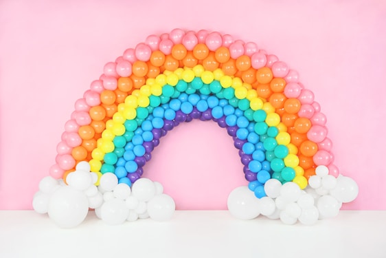 A vibrant arch made of colorful balloons mimicking a rainbow. The arrangement features red, orange, yellow, green, blue, and purple balloons, forming a semicircular shape. At the base of the arch are clusters of white balloons resembling clouds, set against a soft pink background.